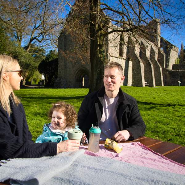 Family enjoying a picnic on a sunny day at a picnic table in front of Grey Abbey, County Down.