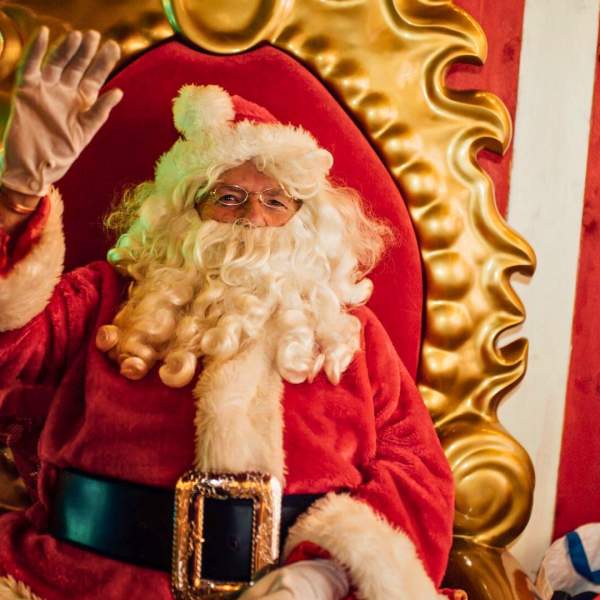 A jolly looking Santa waves as he sits on a plush throne in his grotto