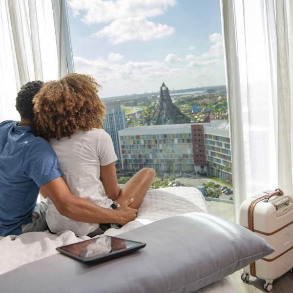 A man and a woman look out the window of their hotel room at Universal Aventura Hotel. They are seen from behind sitting side by side on the bed, cuddling and admiring the striking view of a towering volcano and waterslides at Universal Volcano Bay.