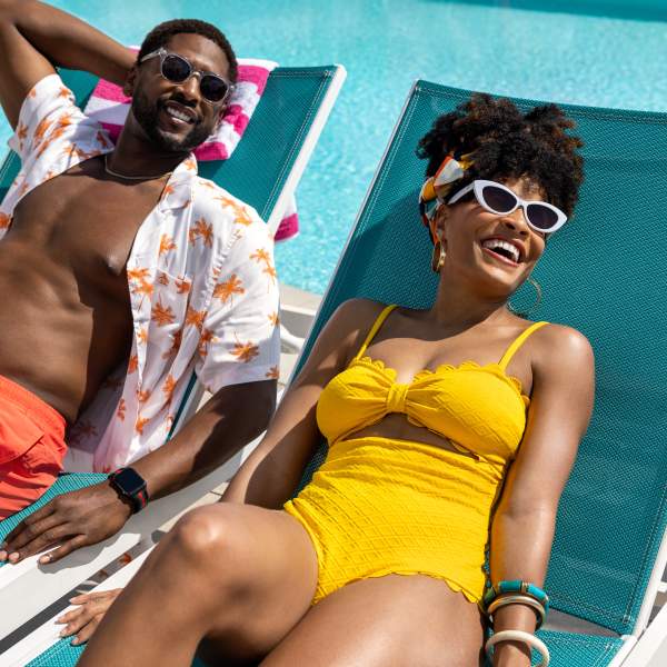 A couple lounge by the pool wearing sunglasses and brightly colored swimsuits