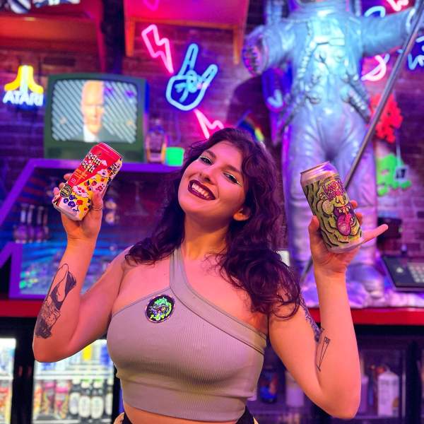 A smiling woman holding two colorful drink cans at 1-Up Orlando, a vibrant, retro-themed bar featuring neon lights, a space-themed statue, and vintage arcade decor in the background.