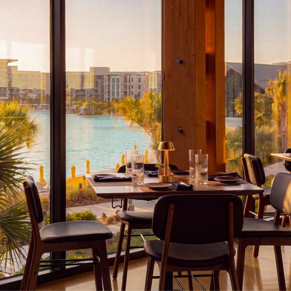 Interior of Twin View Restaurant at Evermore Orlando Resort, with palm trees, a white beach and a shimmering blue bay seen through the floor-to-ceiling window.