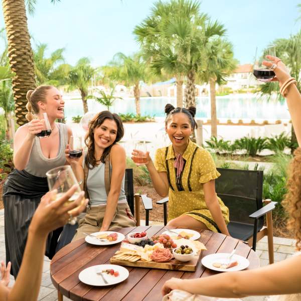 Guests sit around a wooden table with shared snacks and drinks near a tropical lagoon at Evermore Orlando Resort in Orlando, Florida
