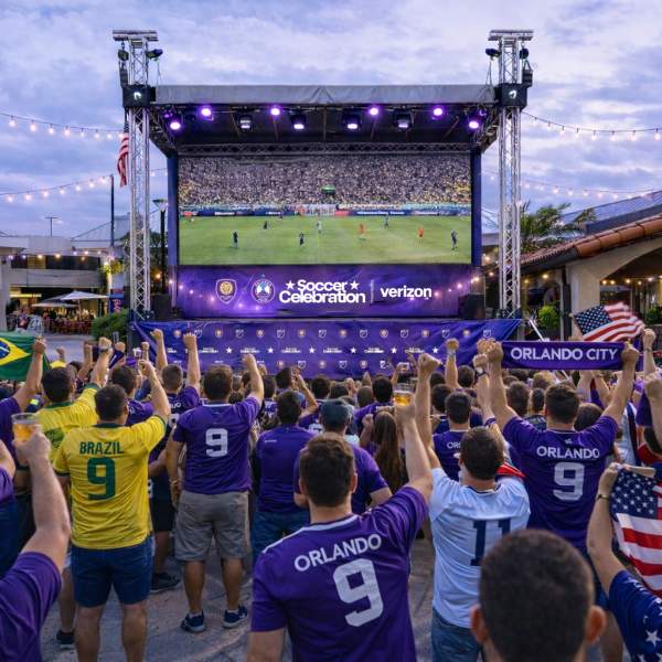 Renderings of viewing areas, Outdoor watch party with a crowd gathered in front of a large elevated screen displaying a soccer match. Attendees wear team jerseys and hold flags while standing beneath string lights and stage lighting in an open-air venue.