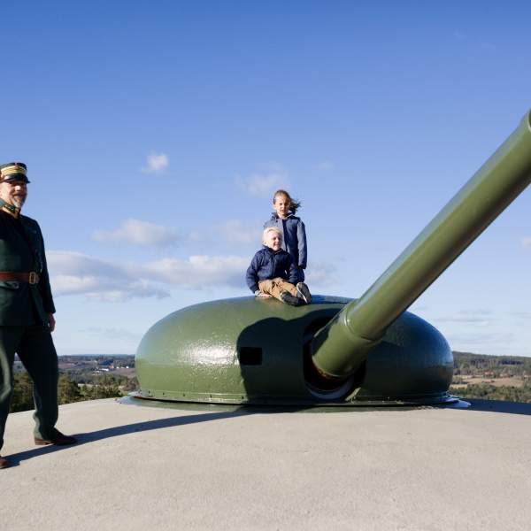 En uniformert mann peker utover ved kanonen på Høytorp fort, to barn sitter på kanonen