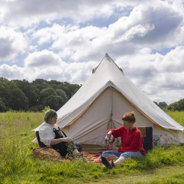 A white canvas tent with two ladies sat in front of it in the grass having a picnic