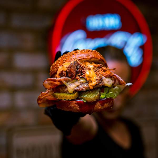 A close up of a man holding a burger up to camera in a dim lit restaurant
