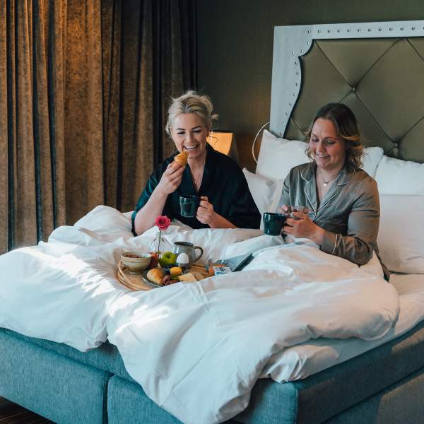 To damer som spise mat og drikke kaffe i en hotellseng. De ler og hygger seg.