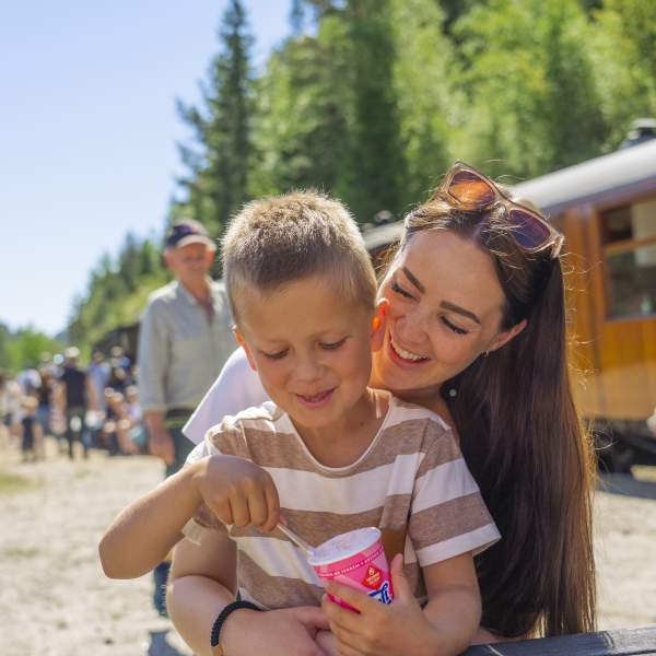Mor og sønn spiser is i sola med gammelt lokmotiv tog i bakgrunnen