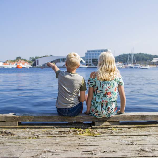 Two children sitting on a dock, pointing towards a building