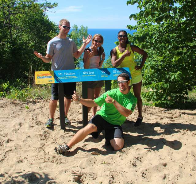 Four athletes stand atop a sand dune near a 3 Dune Challenge sign. The blue water of Lake Michigan is in the distance.