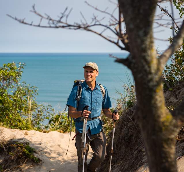 A man in a blue shirt and light cap hikes on a sandy trail with hiking poles. Lake Michigan can be seen in the background.