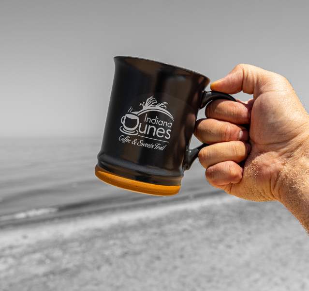 A masculine hand holds a coffee mug in front of a lake and beach. The photo is black and white except for the orange rim on the mug and the hand.