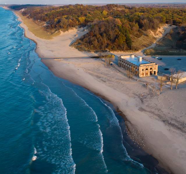 Bright blue water with white cresting waves near the shore sits to the left. On the right is a long sandy beach that spreads to the horizon line. Wooded dunes extend from the beach and into the distance. A stone building sits on the beach with a parking lot behind it.