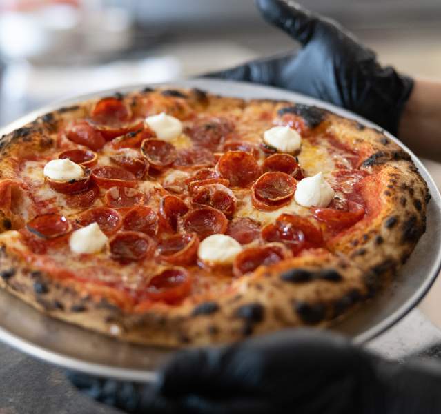 Two hands with black gloves hold a pizza on a metal dish. The pizza has pepperoni and cheese on top.