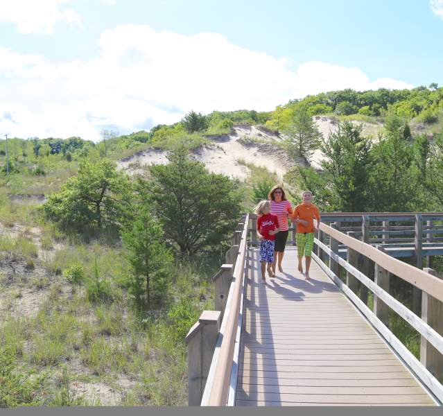 A woman and two small children hike along a wooden boardwalk over vegetation-covered dunes.