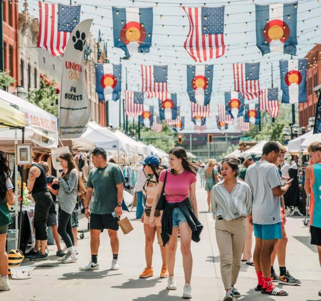 A festival in Larimer Square has American and Colorado flags hanging above as people explore vendors