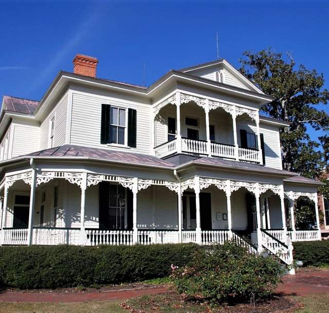 The 1897 Poe House, a two-story white Victorian home in Fayetteville, NC, featuring an ornate wraparound porch, decorative trim, and an upper balcony under a bright blue sky.