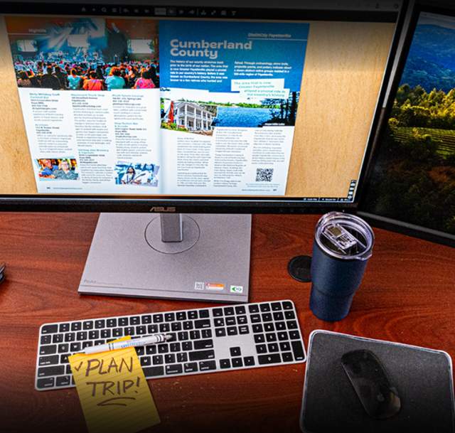 A digital destination guide is open on a computer monitor at an office desk with a keyboard, mousepad, a blue travel cup, and a yellow sticky note that says Plan Trip.