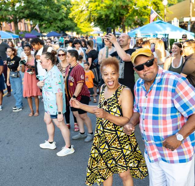 Diverse group of people dancing and smiling at a Fourth Friday event in downtown Fayetteville, NC, with vendor tents and summer sunlight in the background.