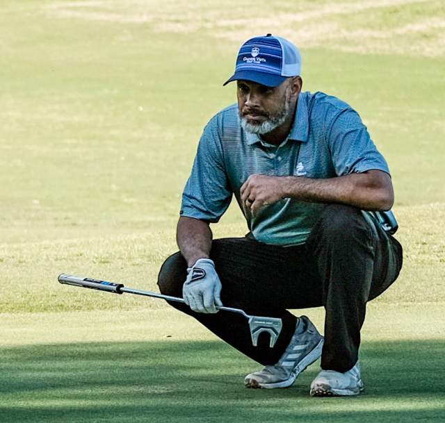 Golfer in a blue shirt and cap crouching on the green while lining up a putt at a local golf course in Cumberland County, NC.