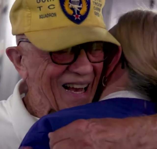 Older military veteran wearing a yellow cap smiles while embracing a woman during an emotional reunion moment, captured in a close-up setting.