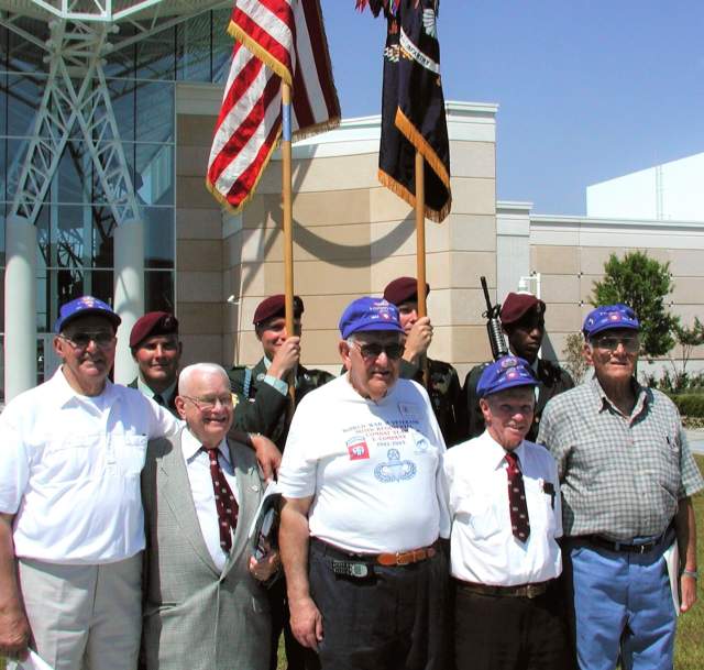 A group of veterans wearing matching caps poses for a photo outside a modern building, with color guard members holding U.S. and military flags behind them.