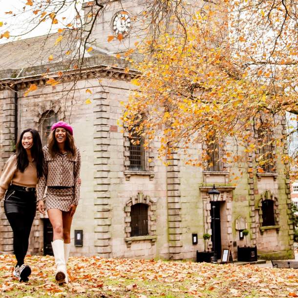 Two friends walking through a park in autumn with fallen orange and red leaves with St Phillip's Cathedral in the background