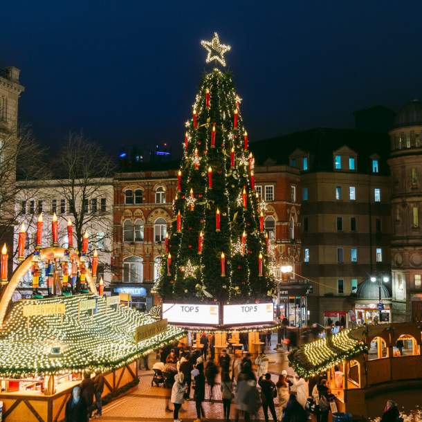 A tall Christmas tree stands in a square in Birmingham at night, the lights are lit and people are gathered round its base at Christmas market stalls