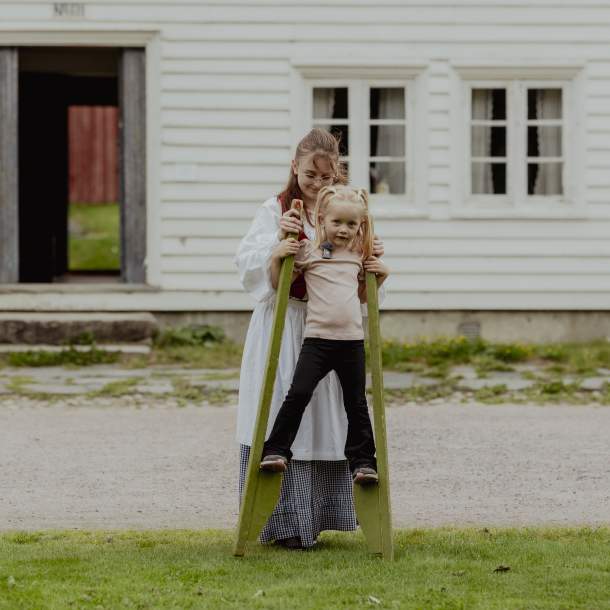 Jente prøver stylter foran gammeldagse hus, mens en kvinne i historiske klær hjelper henne