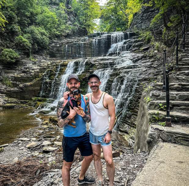 Two men stand in front of a waterfall, holding a small dog.