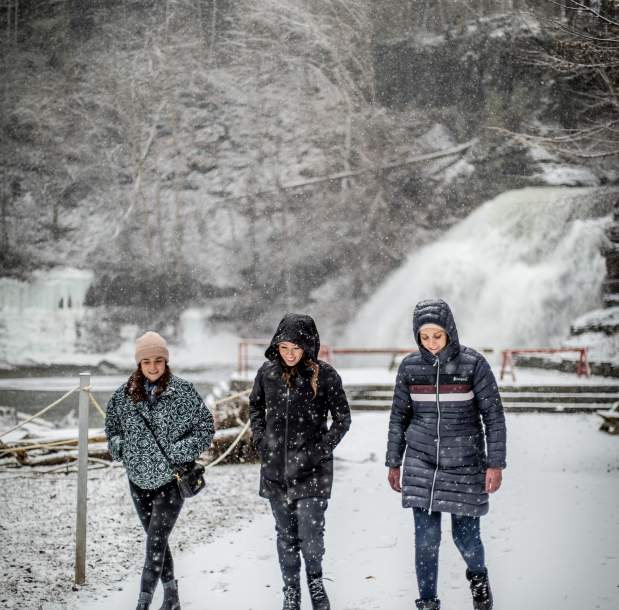 three people in parkas walk near waterfall in winter