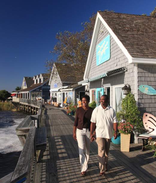 A couple strolls hand-in-hand along a scenic boardwalk by the water, surrounded by colorful shops and vibrant coastal flora.