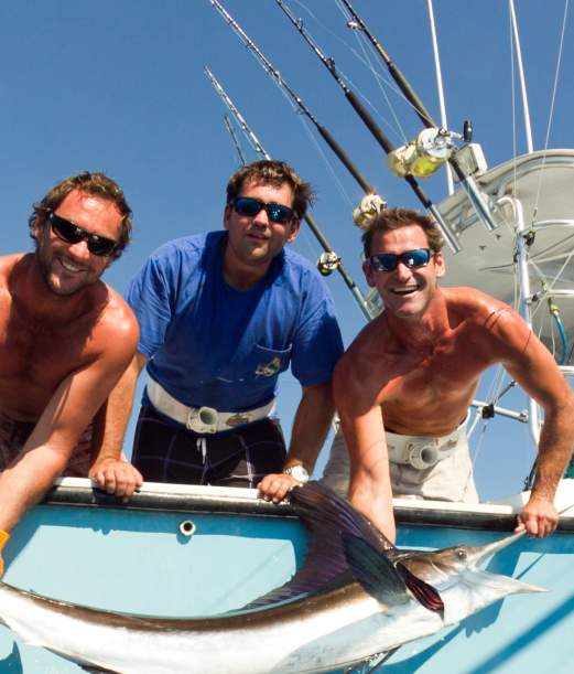 Three men on a fishing boat hold up a large fish, showcasing their catch under a clear blue sky.