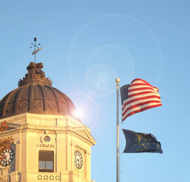 Bloomington courthouse dome with weather vane beside American and Indiana flags waving against a clear blue sky.