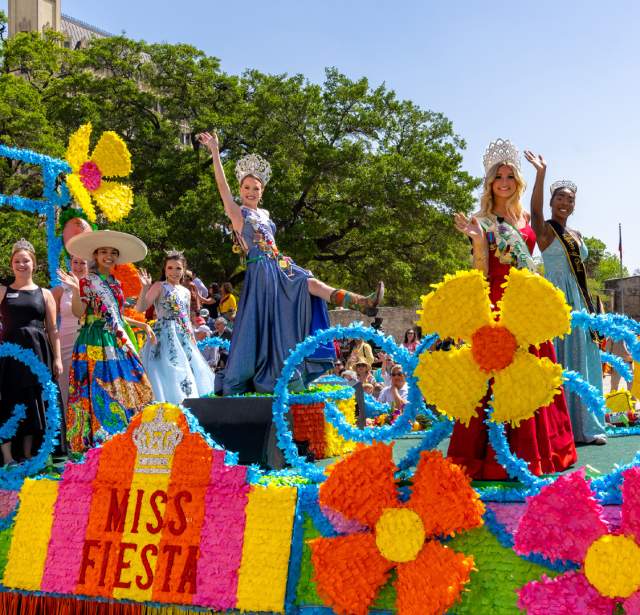 Women dressed in gowns and crowns on a colorful Fiesta float in front of the Alamo.