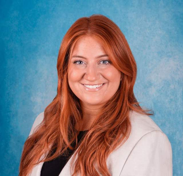 Professional headshot of a woman with red hair in a white blazer and black top.