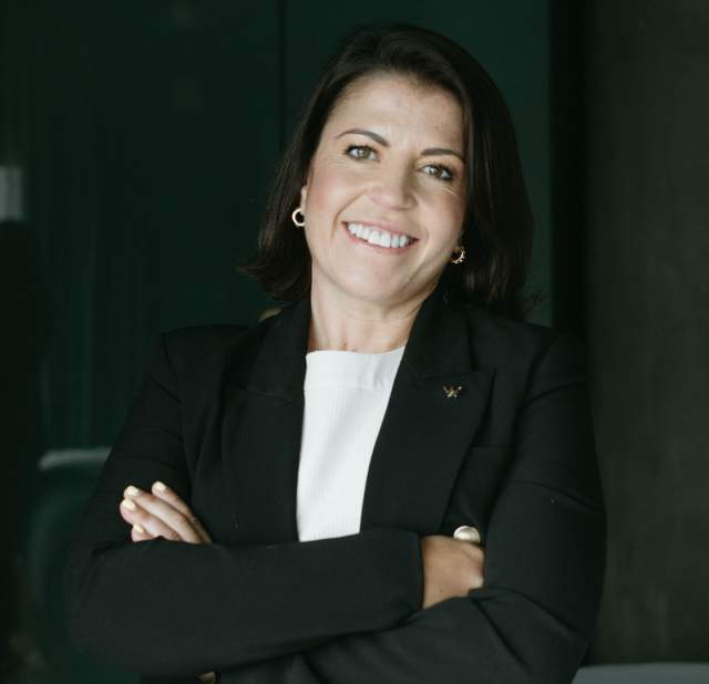 A woman wearing a white blouse and a black jacket, looking directly at the camera and smiling with her arms crossed across her chest.