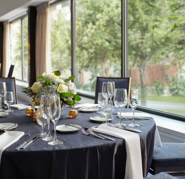An event room table set with a blue tablecloth and white napkins; the room has a view of lush green trees.