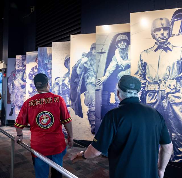 Two visitors walk through a military history exhibit lined with large blue-toned panels of paratroopers and nearby display cases at Fayetteville's ASOM.
