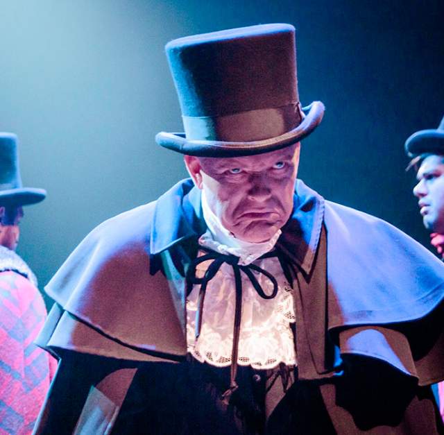 Actors in Victorian costumes and top hats perform A Christmas Carol on stage in Fayetteville, NC, with a stern central figure in a cape under dramatic blue lighting.
