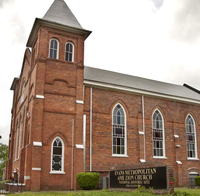 Evans Metropolitan AME Zion Church in Fayetteville—a historic red-brick sanctuary with a pointed steeple and tall arched stained-glass windows, its sign and landscaped lawn visible under a cloudy sky.