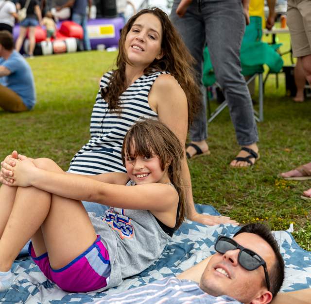 A family relaxes on a picnic blanket at Fayetteville, NC's Greek Festival, with a smiling young girl in the center and inflatable bounce houses in the background.