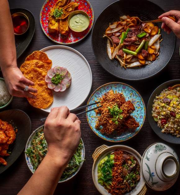 An overhead view shows multiple hands reaching toward a shared selection of plated dishes arranged across a dining table at Yao’s Restaurant. Bowls, plates, and utensils emphasize a communal dining experience.