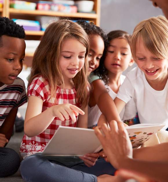 Group Of Elementary School Pupils Sitting On Floor Listening To Female Teacher Read Story