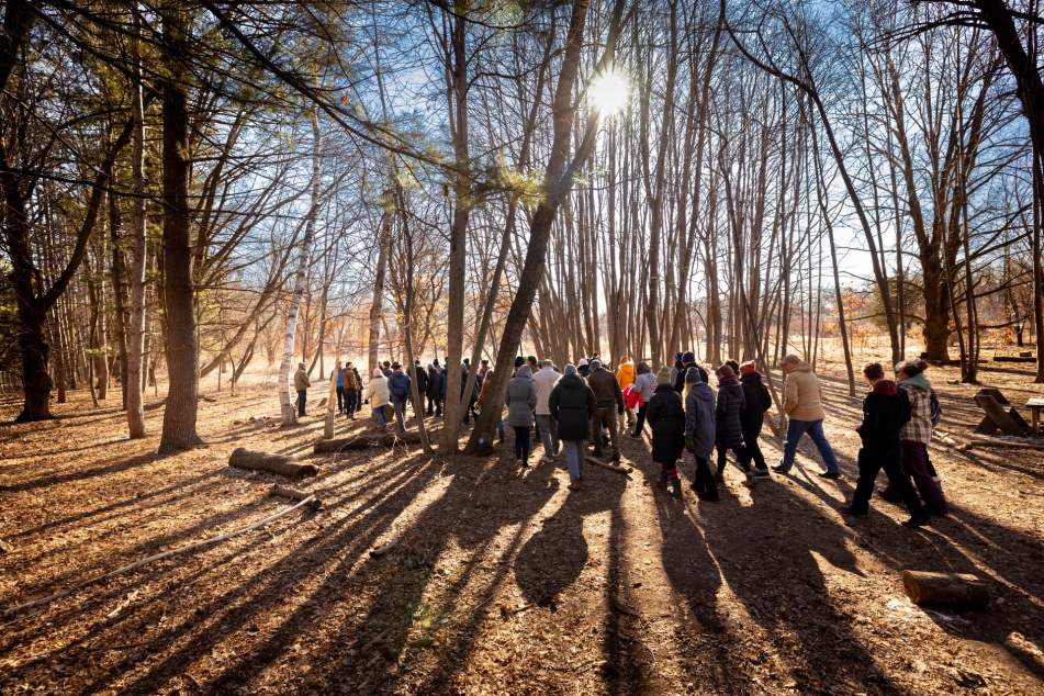 A group of a few dozen people hike in a tall wooded forest on a sunny winter afternoon at the Aldo Leopold Nature Center