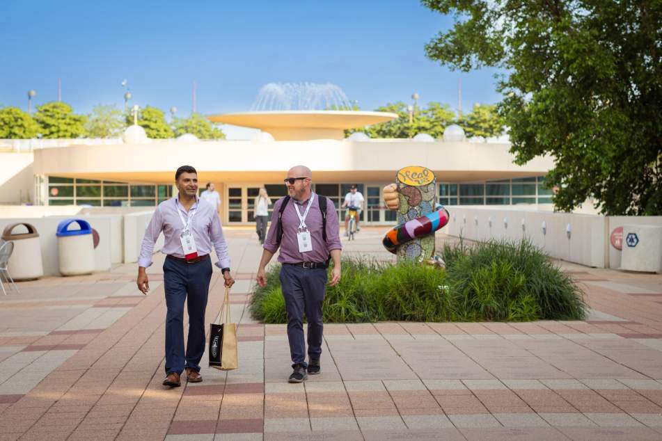Two men dressed in business clothes walk from a STEM conference at the Monona Terrace Community and Convention Center with a fountain in the background.