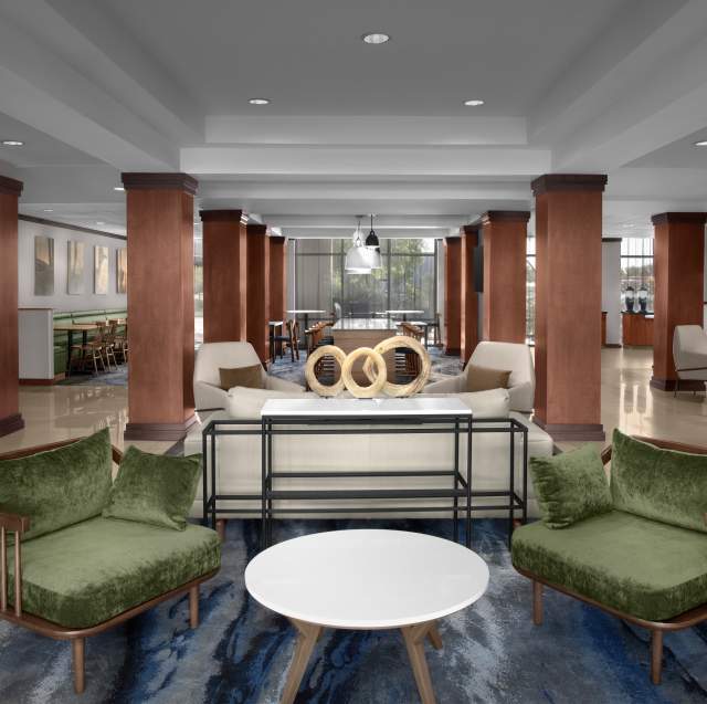 Lobby with cherry wood pillars throughout, a wooden lobby check-in desk to the right and seating in the middle with sandy colored tile floors. The middle of lobby has two velvet green chairs with a small circular side table in the middle, a white entry table with metal framing, and a white couch with two white chairs behind the entry table. A blue modern streaked carpet runs down the middle. In the background a variety of bar stool and cafe-style seating can be seen.