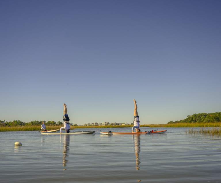 Women practicing paddleboard yoga with headstands on calm marsh waters in North Myrtle Beach, South Carolina, under a clear blue sky surrounded by peaceful natural scenery.