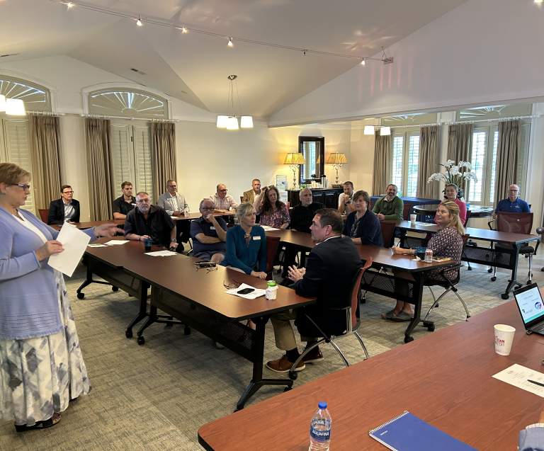 A woman stands at the front of a meeting room holding papers and speaking to a group of seated attendees. People listen attentively around conference tables in a well-lit room with large windows and curtains.